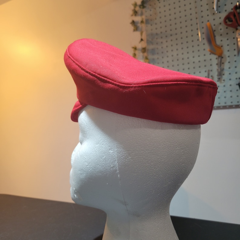 May include: A red fabric hat with a flat top and a curved brim. The hat is on a white mannequin head.