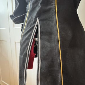 May include: A black coat with a vertical gold stripe and a gray stripe on the side. The coat has a slit on the side, revealing a red garment underneath. The coat is hanging on a white door.