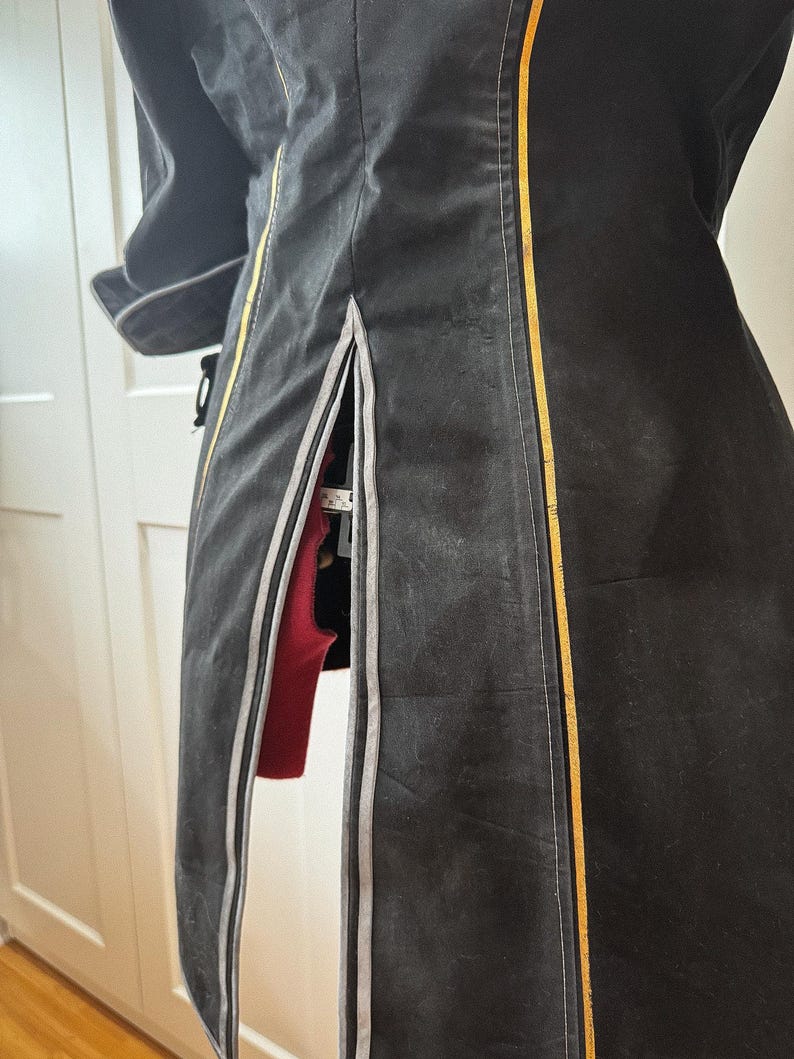 May include: A black coat with a vertical gold stripe and a gray stripe on the side. The coat has a slit on the side, revealing a red garment underneath. The coat is hanging on a white door.