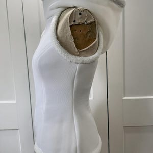 May include: A white hooded vest with a faux fur trim around the hood and hem. The vest is displayed on a mannequin. The hood is lined with the same faux fur. The vest has a fitted silhouette and a smooth, clean texture.