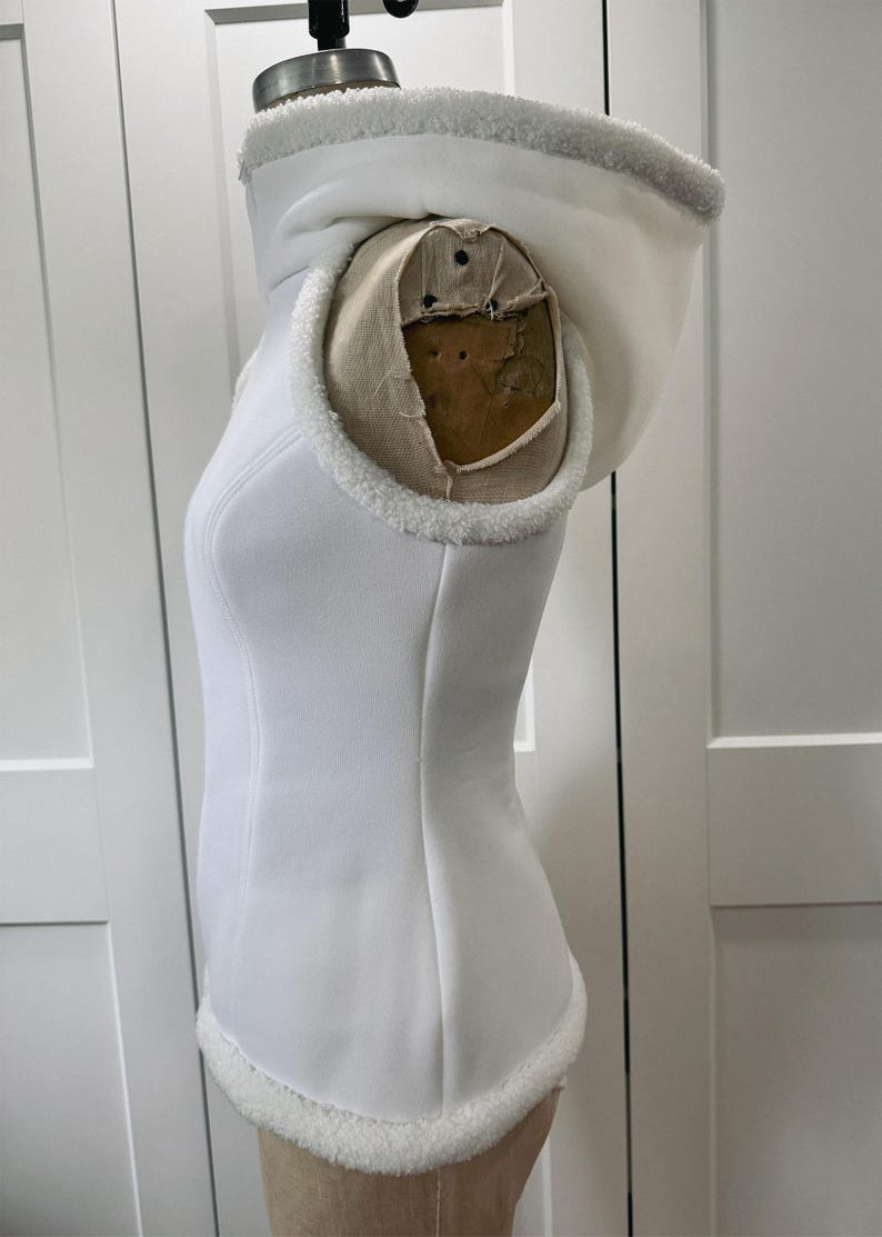 May include: A white hooded vest with a faux fur trim around the hood and hem. The vest is displayed on a mannequin. The hood is lined with the same faux fur. The vest has a fitted silhouette and a smooth, clean texture.