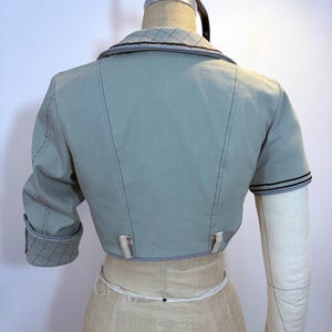 May include: A light blue bolero jacket with short sleeves, displayed on a mannequin. The jacket features a notched collar with a quilted pattern, black stitching, and decorative tabs at the waist. The sleeve cuffs also have a quilted pattern.