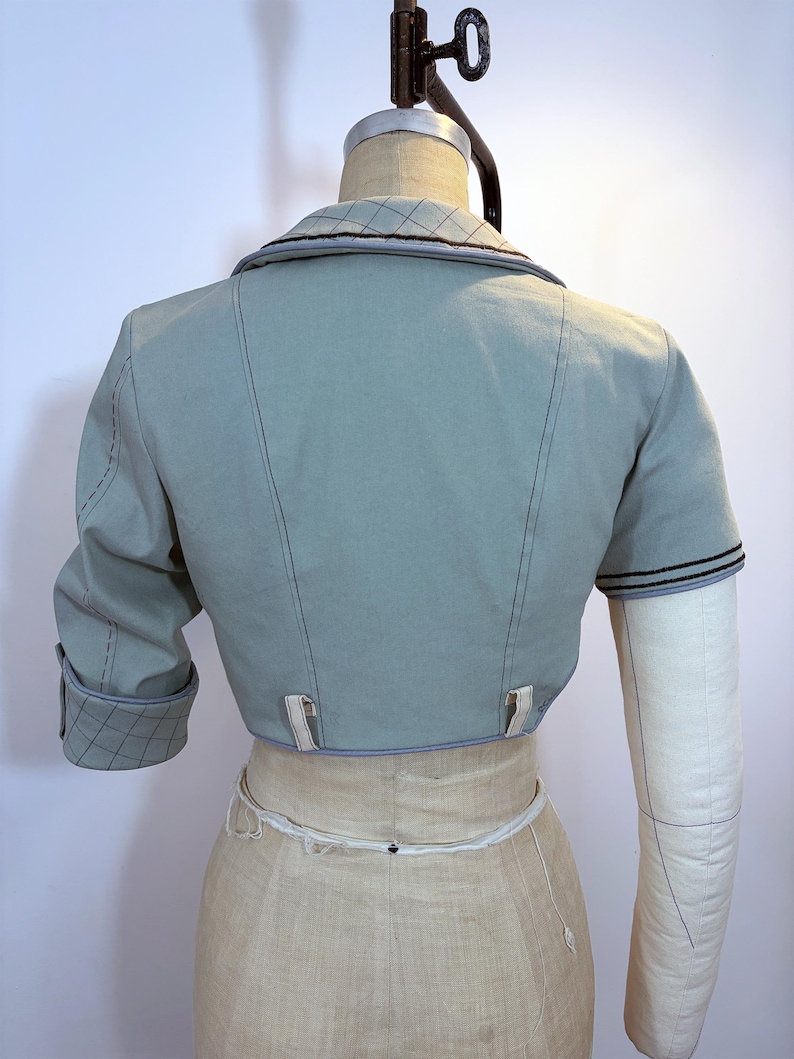 May include: A light blue bolero jacket with short sleeves, displayed on a mannequin. The jacket features a notched collar with a quilted pattern, black stitching, and decorative tabs at the waist. The sleeve cuffs also have a quilted pattern.