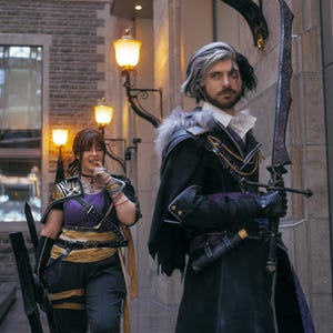 May include: Two people in cosplay costumes pose outdoors. One wears a black and purple outfit with a large sword. The other wears a purple top, black pants, and gold accents, holding a long black prop. The background features a brick building and street lamps.