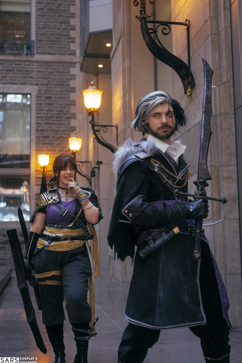 May include: Two people in cosplay costumes pose outdoors. One wears a black and purple outfit with a large sword. The other wears a purple top, black pants, and gold accents, holding a long black prop. The background features a brick building and street lamps.