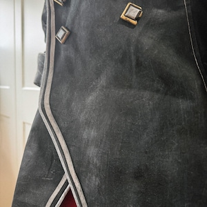 May include: Close-up of a black coat with decorative square buttons and silver trim. The coat has a textured appearance and a red lining visible at the bottom. The buttons are gold-colored with a silver center.