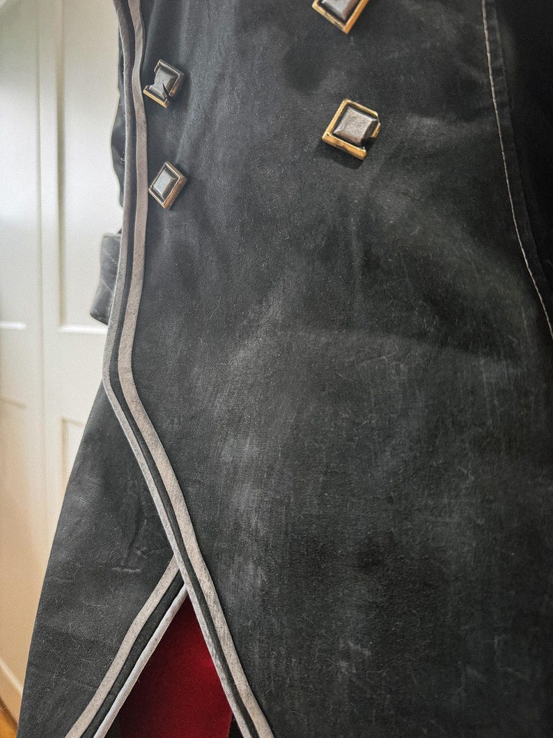 May include: Close-up of a black coat with decorative square buttons and silver trim. The coat has a textured appearance and a red lining visible at the bottom. The buttons are gold-colored with a silver center.
