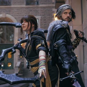May include: Two people in cosplay costumes stand back-to-back, holding weapons. The person on the left has a large, ornate blade, while the person on the right holds a sword and a dagger. Both wear black and gold outfits.