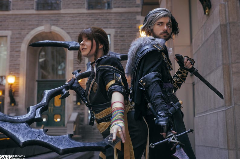 May include: Two people in cosplay costumes stand back-to-back, holding weapons. The person on the left has a large, ornate blade, while the person on the right holds a sword and a dagger. Both wear black and gold outfits.