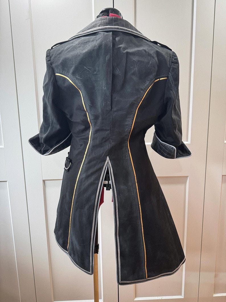 May include: A black coat with gold and silver trim, displayed on a dress form. The coat features a notched collar, short sleeves, and a split back. The design includes decorative stitching and a tailored fit.