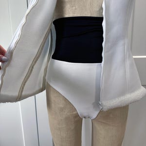 May include: A white bodysuit with a high-cut leg and a black waist shaper. A white zip-up vest with a faux fur trim is partially open, revealing the bodysuit and waist shaper. The outfit is displayed on a mannequin.