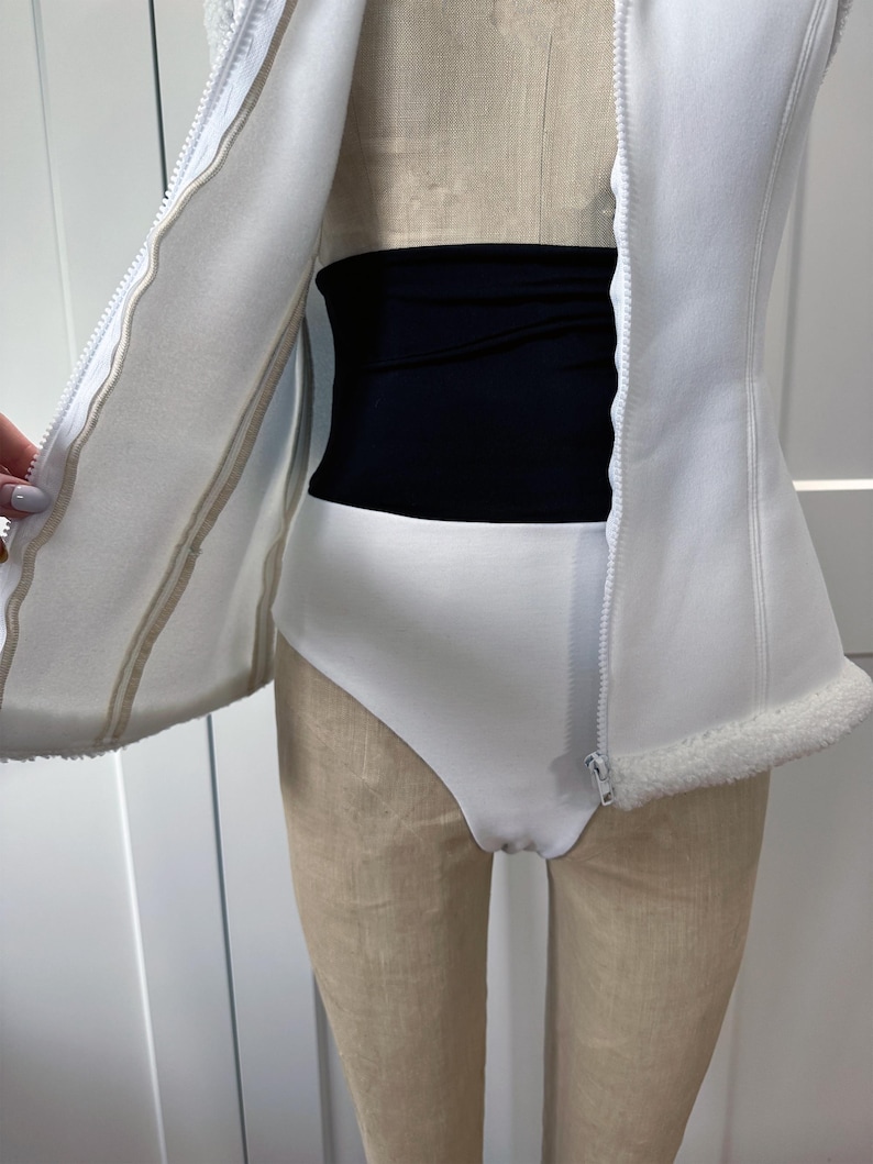 May include: A white bodysuit with a high-cut leg and a black waist shaper. A white zip-up vest with a faux fur trim is partially open, revealing the bodysuit and waist shaper. The outfit is displayed on a mannequin.