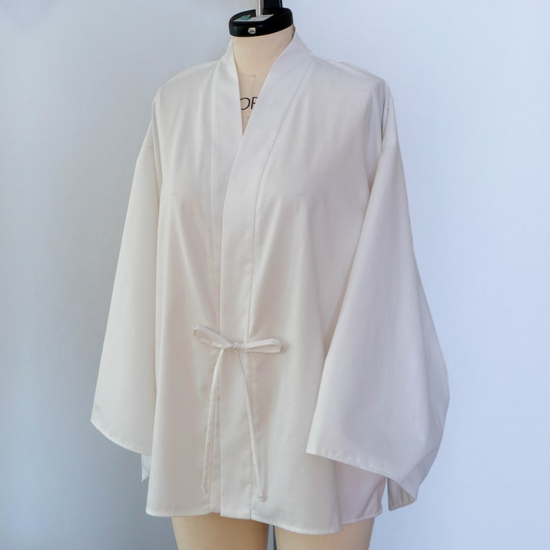 Puede incluir: Un kimono blanco con cierre de lazo. El kimono tiene mangas largas y un corte holgado.
