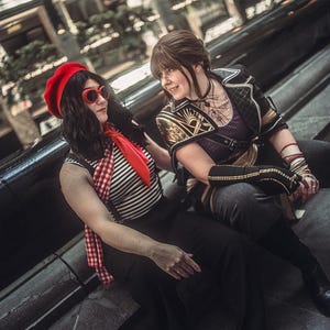 May include: Two people are seated on a dark surface. One wears a red beret, sunglasses, a striped shirt, and a red scarf. The other wears a purple top with gold accents and black pants. The setting appears to be an indoor public space.