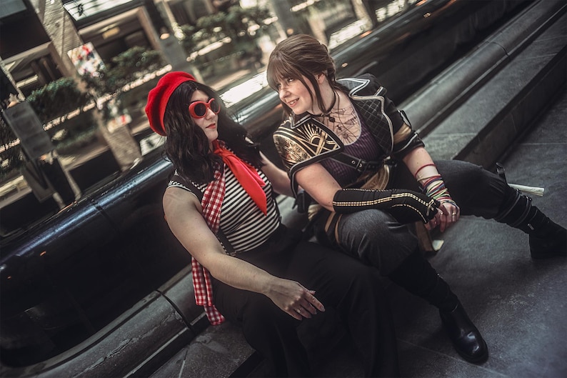 May include: Two people are seated on a dark surface. One wears a red beret, sunglasses, a striped shirt, and a red scarf. The other wears a purple top with gold accents and black pants. The setting appears to be an indoor public space.