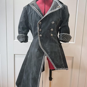 May include: A black coat with a unique design, featuring a double-breasted front with silver buttons and trim. The coat has a long, asymmetrical hemline and quilted detailing on the collar and cuffs. The coat is displayed on a red dress form.