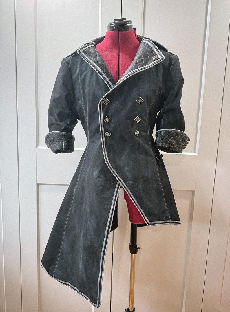 May include: A black coat with a unique design, featuring a double-breasted front with silver buttons and trim. The coat has a long, asymmetrical hemline and quilted detailing on the collar and cuffs. The coat is displayed on a red dress form.