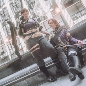 May include: Two people in cosplay outfits, one standing and one seated. The standing person wears a purple top, black pants, and a belt. The seated person wears a purple jacket and black pants. Both have black boots. The background is a city scene.
