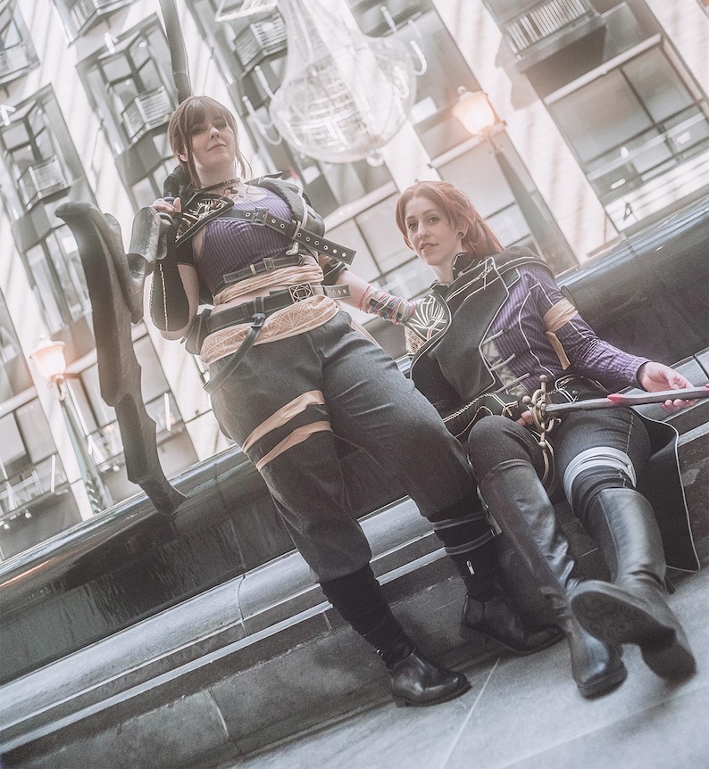 May include: Two people in cosplay outfits, one standing and one seated. The standing person wears a purple top, black pants, and a belt. The seated person wears a purple jacket and black pants. Both have black boots. The background is a city scene.