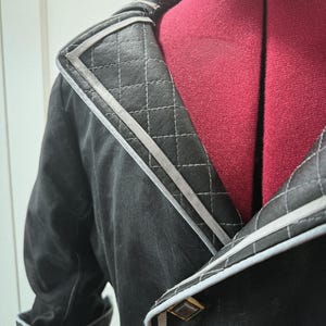 May include: Close-up of a black jacket with a quilted collar and cuffs. The jacket features gray trim and gold-colored buttons. The collar has a diamond-stitched pattern. The jacket is displayed on a red dress form.