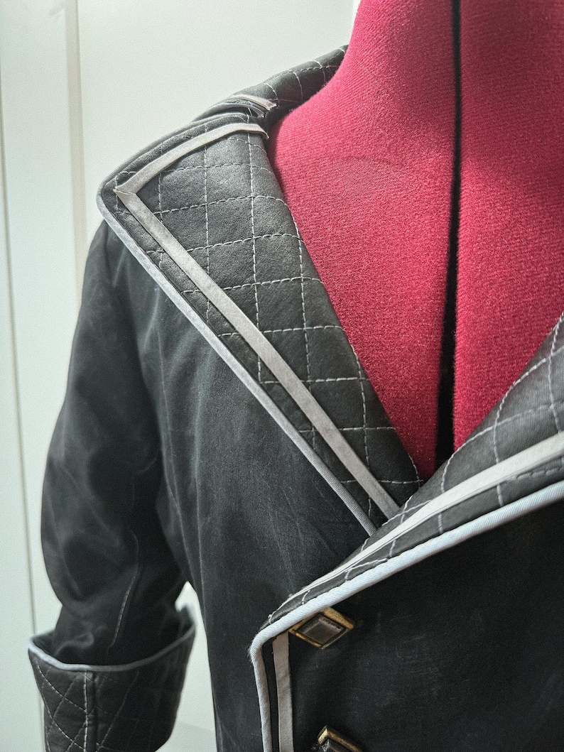 May include: Close-up of a black jacket with a quilted collar and cuffs. The jacket features gray trim and gold-colored buttons. The collar has a diamond-stitched pattern. The jacket is displayed on a red dress form.