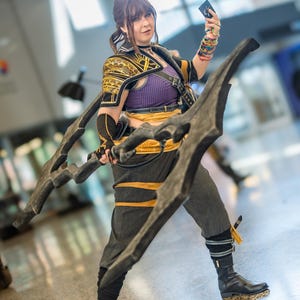 May include: A person in a cosplay costume, holding a large, dark-colored weapon. The costume features a purple top, black and gold accents, and gray pants. The person is also holding a small card.