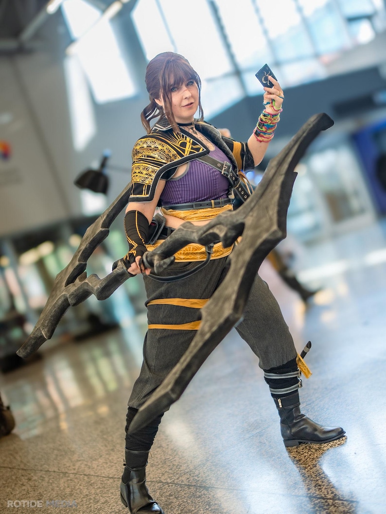 May include: A person in a cosplay costume, holding a large, dark-colored weapon. The costume features a purple top, black and gold accents, and gray pants. The person is also holding a small card.