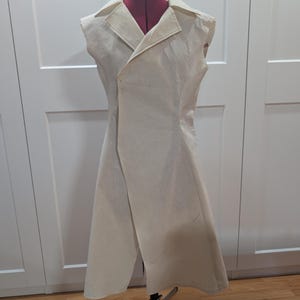 May include: A cream-colored, sleeveless coat displayed on a red dress form. The coat features a notched lapel, a single-button closure, and a wrap-style design. The garment is made of a light-colored fabric and has a tailored fit.