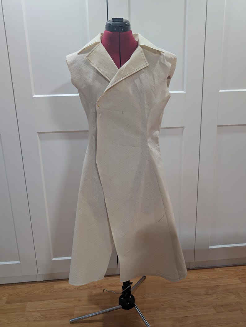 May include: A cream-colored, sleeveless coat displayed on a red dress form. The coat features a notched lapel, a single-button closure, and a wrap-style design. The garment is made of a light-colored fabric and has a tailored fit.