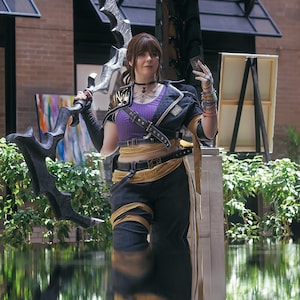 May include: A person in a cosplay outfit, holding a large, ornate weapon. The costume includes a purple top, black jacket, and dark pants with gold accents. The person is standing near water, with greenery and artwork in the background.