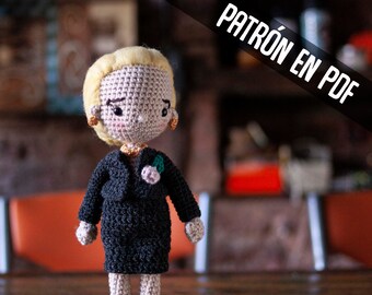 May include: A crocheted doll wearing a black suit and a small pink flower. The doll has blonde hair and a light pink face. The doll is standing on a wooden surface. The text "PATRÓN EN PDF" is visible in the background.