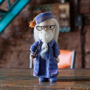 May include: A crocheted doll wearing a blue robe, a brown hat with orange trim, and glasses. The doll has a white beard and is holding a brown stick.