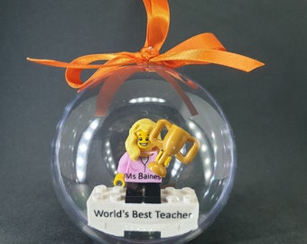 Puede incluir: Un adorno esférico transparente que contiene una minifigura rubia con una camisa rosa etiquetada como Ms Baines y sosteniendo un trofeo dorado. La figura se apoya sobre una base de ladrillo blanco con el texto World's Best Teacher y un lazo naranja.