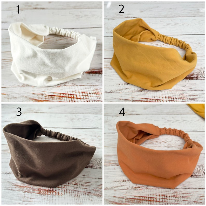 May include: A set of four wide fabric headbands featuring an elasticized back strap for a secure fit. The collection includes one white, one mustard yellow, one dark brown, and one terracotta orange headband, each with a smooth, solid-colored finish.