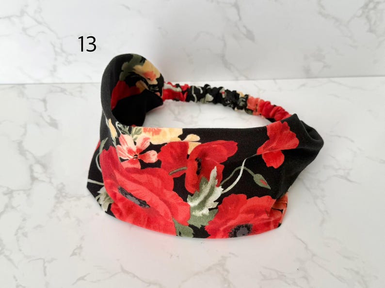 May include: A fabric headband featuring a black background with a vibrant floral pattern of large red poppies, smaller yellow flowers, and green leaves. The accessory includes a gathered elastic band section at the back for a secure fit.