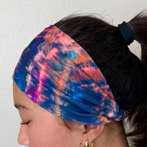May include: A wide fabric headband featuring a vibrant tie-dye pattern with blended streaks of deep blue, bright pink, purple, and orange. The material is gathered at the sides and fits snugly around the head, covering the hairline.