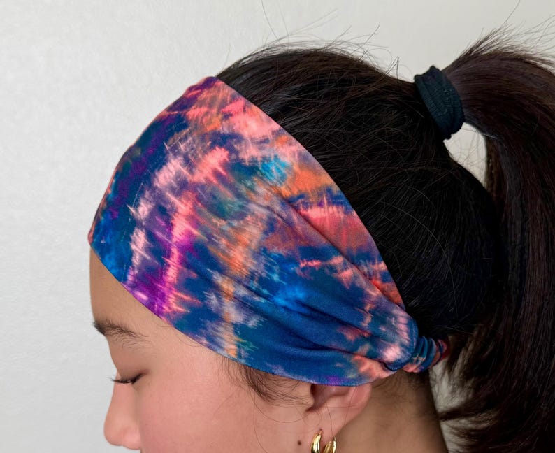 May include: A wide fabric headband featuring a vibrant tie-dye pattern with blended streaks of deep blue, bright pink, purple, and orange. The material is gathered at the sides and fits snugly around the head, covering the hairline.