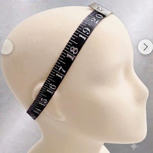 May include: A black measuring tape labeled Premium Head Circumference Guide wrapped around a beige mannequin head. The tape features white numerical markings from 14 to 20 inches, with a silver metal clasp at the top displaying a crown logo.