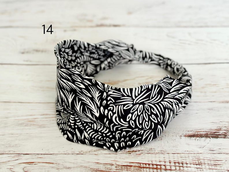 May include: A black and white headband featuring a repeating abstract botanical pattern with a twisted knot detail at the top. The number 14 is printed in the upper left corner of the image. The fabric has a wide, gathered design.