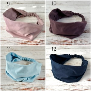 May include: A collection of four wide fabric headbands featuring a gathered elastic back for a secure fit. The set includes one dusty rose, one deep plum, one light sky blue, and one dark navy blue headband, each with a smooth, matte finish.