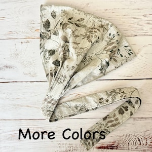 May include: A triangular head wrap featuring a light beige fabric base with a dark gray botanical print of leaves and stems. The head covering includes a wide band and long ties for securing, with the text More Colors printed at the bottom.