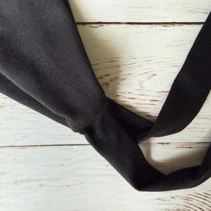 May include: A solid black fabric headband featuring a twisted knot detail at the center. The headband is constructed from a soft, matte material and consists of a wide band that tapers into a knotted section, creating a gathered appearance.