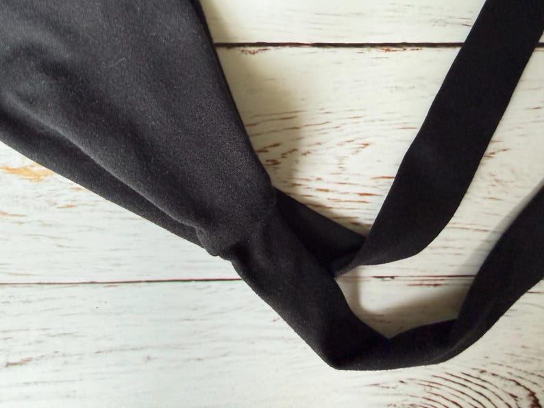 May include: A solid black fabric headband featuring a twisted knot detail at the center. The headband is constructed from a soft, matte material and consists of a wide band that tapers into a knotted section, creating a gathered appearance.