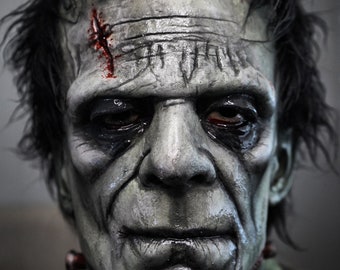 May include: A detailed close-up of a Frankenstein monster mask. The mask is painted in shades of gray and green, with visible stitches, scars, and dark eye sockets. The hair is black and messy, and the overall effect is a realistic, horror-themed prop.