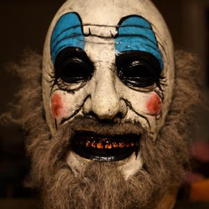 Puede incluir: Una máscara de payaso espeluznante con una cara blanca, cejas azules y maquillaje de ojos negro. La máscara tiene una gran sonrisa inquietante con dientes oscuros y una barba marrón. Rubor rojo en las mejillas.