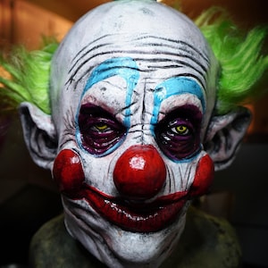 Könnte beinhalten: Eine gruselige Clownsmaske mit weißem Gesicht, grünen Haaren, blau-weißem Make-up, einer roten Nase und einem breiten, zahnigen Grinsen.