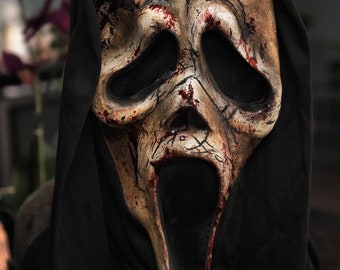 May include: A Scream mask with a distressed, aged appearance. The mask is off-white with dark eye sockets and a gaping mouth. Red paint simulates blood splatters. The mask is framed by a black hood, creating a spooky Halloween costume.