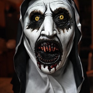 Könnte beinhalten: Nahaufnahme einer Horror-Maske, die eine dämonische Nonne darstellt. Die Maske hat ein blasses Gesicht mit schwarzem Augen-Make-up, gelben Augen und einem klaffenden Mund mit scharfen Zähnen und Blut. Eine schwarze Kapuze umrahmt das Gesicht.