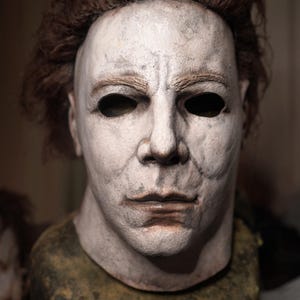 May include: A white mask with black eye holes and a brown wig. The mask has a painted-on face with a neutral expression. The mask is on a green and brown stand.