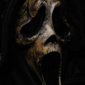 May include: A Scream mask with a distressed, aged appearance. The mask is primarily off-white with areas of gold and black, and features large eye openings and an open mouth. The mask is set against a black background.